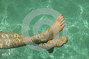 Child`s feet in the blue transparent water of the pool.