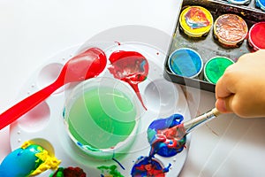 Child`s hand holding brush dipped in paint on white paper background with various type gouache watercolor tempera. Kids creativity