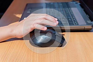 A child`s hand holds a computer mouse next to a laptop keyboard on a wooden table