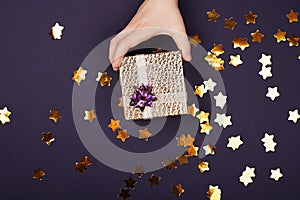 A child s hand holds a gold gift box with a leather-like texture on a purple background with sparkles in the shape of