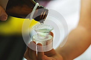 A child`s hand holds a measuring cup for cough syrup close-up. The mother pours medicine or antipyretic from a bottle into a spoon