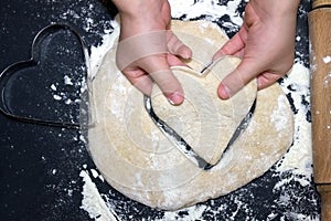 Child`s hands taking a cutted dough heart. Little child preparing dough for backing. A photo from above of Kid`s hands, some flo