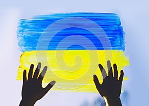 Child`s hands touch painting yellow-blue flag of Ukraine on window. Hands of little kid on image of flag of Ukraine on glass.