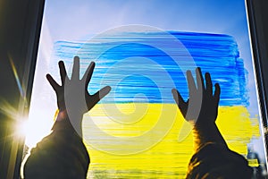 Child`s hands touch painting yellow-blue flag of Ukraine on window. Hands of little kid on image of flag of Ukraine on glass.