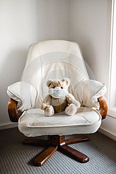 A child`s teddy bear sitting on a chair wearing a face mask