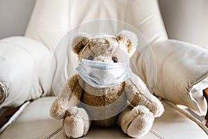 A child`s teddy bear sitting on a chair wearing a facemask