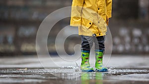 The child`s yellow raincoat and green boots fall into a puddle of water. Autumn rainy weather. A walk in the rain.