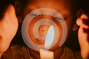 Child with sad eyes looks at flame of burning candle in dark. Memories or mourning for dead. Religious sacrament in church