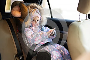 Child safety seat chair baby girl with mobile phone at hands is on back seat of car