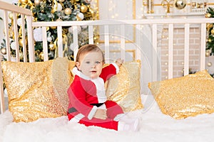 A child in a Santa costume at the Christmas tree with a gold decoration is sitting in a crib at home