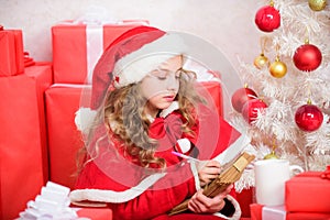 Child santa costume enjoy christmas eve. Girl little kid hold pen and paper near christmas tree writing letter. Child