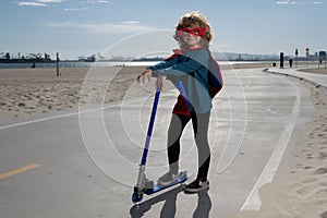 Child on scooter in park. Kids learn to skate roller board. Little boy superhero with hero cloak skating on road