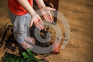 Child showing at dirty soil hands sitting on the floor with a broken pot of flowerpot