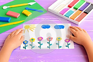 Child shows a card with plasticine flowers, sun and clouds. Supplies for children art crafts on wooden table. Modeling clay craft