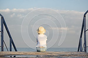 Child sits alone on pier on sea and sky background. Childrens loneliness