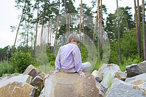 the child sits on the bark of large stones in front of a pine forest.