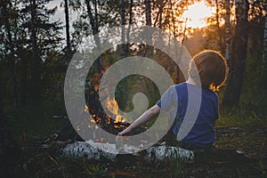 A child sits in a forest near a fire and looks at the setting sun