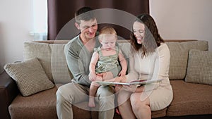 Child sits in his dad arms, his mom reads a book to toddler, looking at educational pictures. Parents spend time with