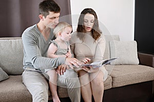 Child sits in his dad arms, his mom reads a book to toddler, looking at educational pictures. Parents spend time with