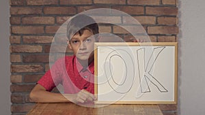 Child sitting at the desk holding flipchart with lettering ok on the background red brick wall.