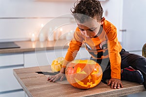 Child sitting at kitchen table dressed as pumpkin playing with decorated halloween pumpkin. Happy Halloween