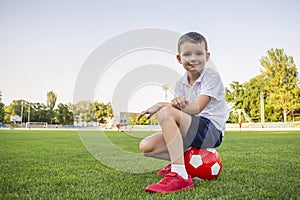 A child is sitting on a soccer ball on the grass of stadium`s field. Training and sports for children concept