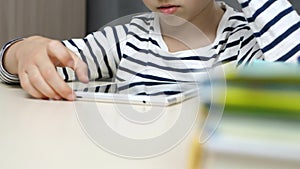 The child is sitting at the table and reading a book using a tablet. The boy is playing, using the app for children. The