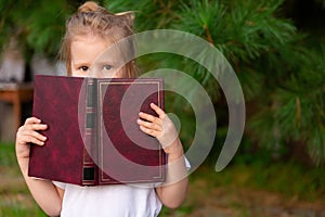 Child slyly narrowed his eyes, holds book. Little girl hiding behind book, looking at camera. Schoolgirl reading book