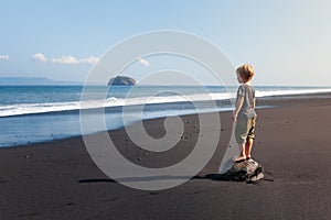 Child stand on big stone on black sand sea beach