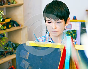 Child standing next to the easel. Kid boy learn paint by brush in class school. Kindergarten interior on background. Boy
