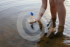 child, standing in a river, launches a home-made boat made of wine corks, instead of sail, a yellow-blue heart