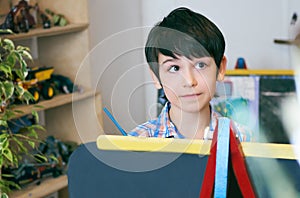 Child standingnext to the easel. Kid boy learn paint by brush in class school. Kindergarten interior on background. Boy