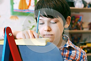 Child standingnext to the easel. Kid boy learn paint by brush in class school. Kindergarten interior on background. Boy