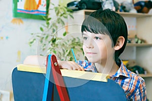 Child standingnext to the easel. Kid boy learn paint by brush in class school. Kindergarten interior on background. Boy