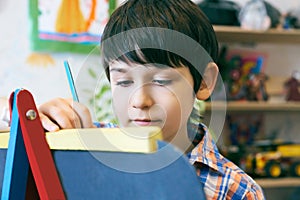 Child standingnext to the easel. Kid boy learn paint by brush in class school. Kindergarten interior on background. Boy