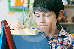 Child standingnext to the easel. Kid boy learn paint by brush in class school. Kindergarten interior on background. Boy