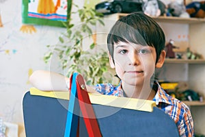 Child standingnext to the easel. Kid boy learn paint by brush in class school. Kindergarten interior on background. Boy