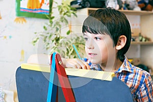 Child standingnext to the easel. Kid boy learn paint by brush in class school. Kindergarten interior on background. Boy