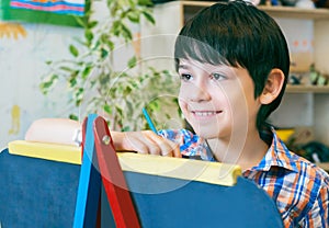 Child standingnext to the easel. Kid boy learn paint by brush in class school. Kindergarten interior on background. Boy