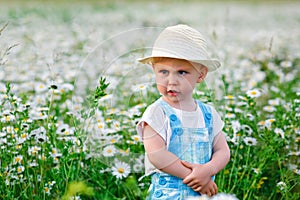 A child in a straw hat in daisy flowers in a field in summer at sunset. A blue-eyed and fair-haired disgruntled boy stands in a