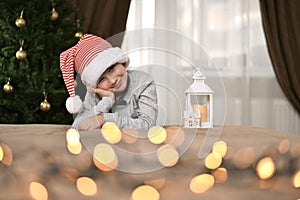 A child in a striped cap at the Christmas tree, with cunning eyes and a kind modest smile, with his cheek propped up.