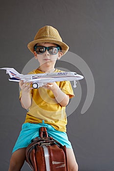 A child in summer clothes and sunglasses sits on a suitcase and holds a toy airplane in his hands. Waiting for travel