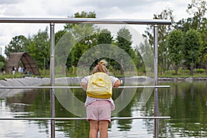 A child in the summer on the pond leaned over the railing and looks at the water. The child looks at the fish swimming