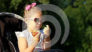 Child in sunglasses is sitting in a stroller and eats an apple at sunset. Portrait baby girl close-up on a sunny day.