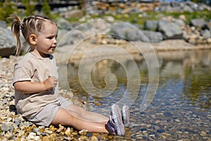 A child in sunny weather is resting on a rocky shore of a lake. A little cute girl with ponytails put her feet in the