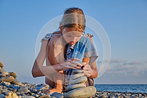 A child in a swimming suit on the seashore is playing with pebbles and building a tower of stones.