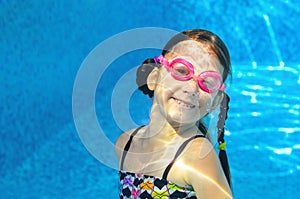 Child swims in pool under water, funny happy girl in goggles has fun and makes bubbles, kid sport