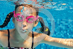 Child swims in pool underwater, funny happy girl in goggles has fun under water and makes bubbles, kid sport