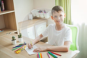The child at the table draws on a blank sheet, around many colored pencils and felt-tip pens. The boy looks at the camera and