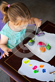 Child in a teal shirt painting rocks colorful colors, art activity for a young toddler or older child